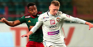 На изображении два футболиста в борьбе за мяч на поле. Игрок в белой форме обходит соперника в зеленой форме. Обстановка матча напряженная, на заднем плане видны размытые трибуны стадиона