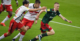 На изображении несколько футболистов в движении на поле. Один игрок в форме с бело-красными элементами пытается догнать другого в зелёно-красной форме. Они сосредоточены на игре. Видна часть стадиона